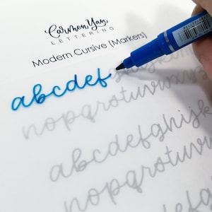 May include: A close-up of a hand using a blue marker to write the letters "abcdef" in cursive on a white sheet of paper. The letters are written over a faint gray outline of the alphabet. The paper has the text "Carmen Yan LETTERING Modern Cursive (Markers)" at the top.