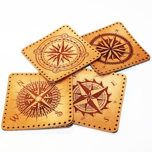 Peut inclure: Quatre sous-verres en cuir marron avec des motifs de boussole. Chaque sous-verre présente une rose des vents différente avec les points cardinaux marqués.