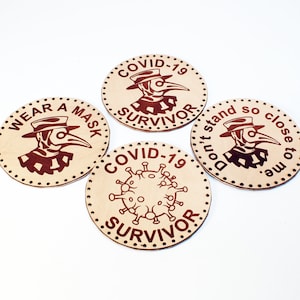 May include: Four round, light brown coasters with dark brown engraved designs. Each coaster features a plague doctor illustration and text: "Wear a Mask", "COVID-19 Survivor", "Don't stand so close to me", and "COVID-19 Survivor".