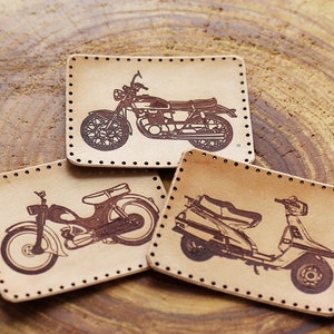 Puede incluir: Tres parches de cuero con bordes cosidos, cada uno con un diseño de motocicleta o scooter vintage. Los parches son marrones y tienen un aspecto desgastado.