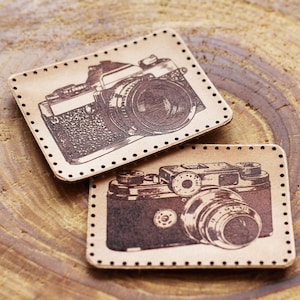 Könnte beinhalten: Zwei Lederpatches mit Vintage-Kamera-Designs. Die Patches sind braun mit schwarzen Nähten und detaillierten Illustrationen von Kameras.
