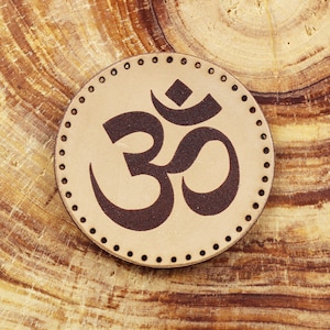 May include: A brown leather patch with a brown Om symbol in the center. The patch has a stitched border with small holes around the edge.