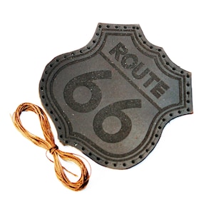 Puede incluir: Un parche de cuero marrón con el texto "ROUTE 66" en letras negras. El parche tiene un borde cosido y se acompaña de un trozo de cuerda marrón.