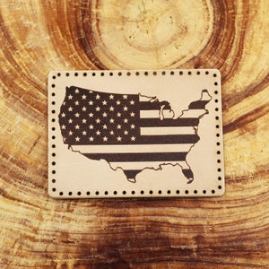 Könnte beinhalten: Ein braunes Lederstück mit einem genähten Rand, das eine schwarz-weiße Umrandung der Karte der Vereinigten Staaten mit dem Design der amerikanischen Flagge im Inneren zeigt.