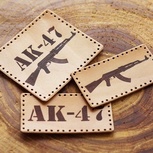 Könnte beinhalten: Drei hellbraune Lederaufnäher mit einem dunkelbraunen AK-47-Gewehrdesign und dem Text "AK-47". Die Aufnäher sind rechteckig und rautenförmig, mit kleinen Löchern an den Rändern.