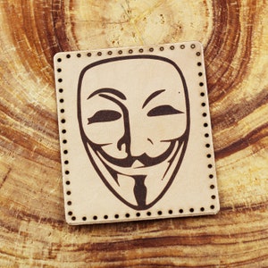 Puede incluir: Un parche de cuero beige con un gráfico en blanco y negro de la máscara de Anonymous. La máscara tiene una sonrisa estilizada y una barba negra.