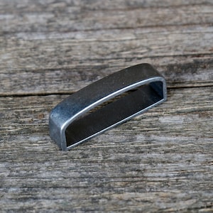Può includere: Una fibbia metallica rettangolare, aperta alle estremità, con finitura argento opaco. La fibbia è posta su una superficie di legno grigio e invecchiato, evidenziando il suo design industriale e l'estetica minimalista. La forma della fibbia suggerisce una cintura o un cinturino.