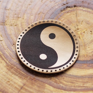 Puede incluir: Un parche redondo de cuero con un símbolo yin yang en blanco y negro en el centro. El parche tiene un borde decorativo de pequeños círculos.