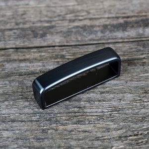 Può includere: Una fibbia rettangolare in metallo nero con bordi arrotondati. La fibbia è posizionata su una superficie di legno grigio e invecchiato, mostrando il suo design elegante e la finitura metallica. La forma della fibbia è un semplice rettangolo.