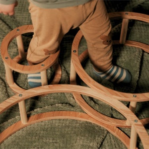 Könnte beinhalten: Ein Kind steht auf einer Holzspielstruktur mit drei runden Ringen. Die Ringe sind aus Naturholz gefertigt und haben eine glatte Oberfläche. Das Kind trägt eine braune Hose und gestreifte Socken.