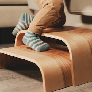 May include: A two-step wooden stool with a curved design. The stool is made of light-colored wood with visible grain. A child's feet, wearing blue and white striped socks, are on the stool. The child is wearing tan pants.