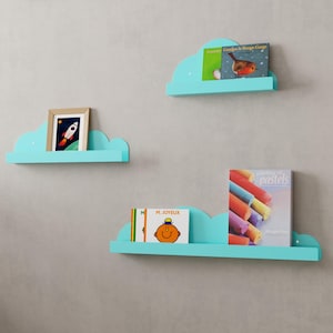 May include: Three turquoise cloud-shaped shelves mounted on a wall. The top shelf has a book titled "Goodbye to Rouge Gorge" on it. The middle shelf has a framed picture of a rocket ship. The bottom shelf has two books, one titled "M. Joyeux" and the other titled "Drawing pastels".