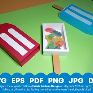 May include: Three popsicle shaped boxes, one red, one blue, and one white with a stick. The white box contains colorful gummy candies. The boxes are on a green background.