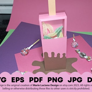 May include: A pink and brown paper craft box shaped like an ice cream cone with a brown chocolate drip. The box is filled with green and yellow gummy candies. The box is sitting on a pink surface with a brown chocolate drip around it.