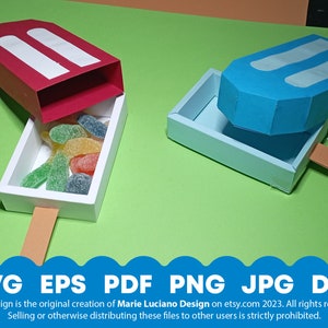 May include: Two open popsicle-shaped boxes, one red and one blue, with white stripes. The red box is filled with colorful gummy candies. The boxes are made of paper and have a stick attached to the bottom.