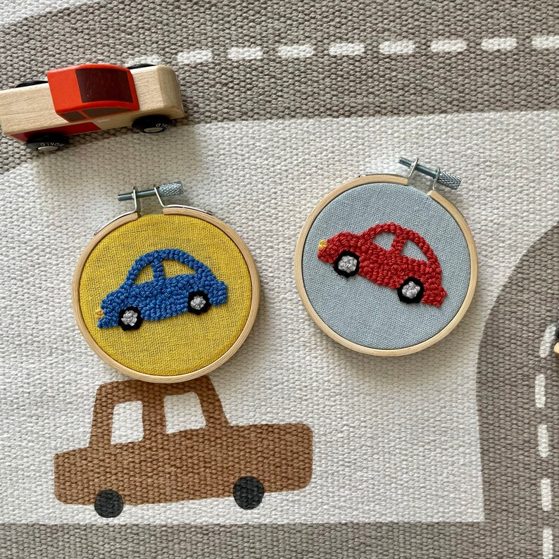 May include: Two embroidery hoops with colorful cars stitched inside. The hoops are on a gray and white patterned surface. One hoop has a blue car and a yellow background. The other hoop has a red car and a light blue background.