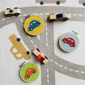 May include: Five wooden toy cars on a gray and white rug with a road design. Four of the cars are in embroidery hoops with colorful yarn. The cars are blue, red, orange, and red.