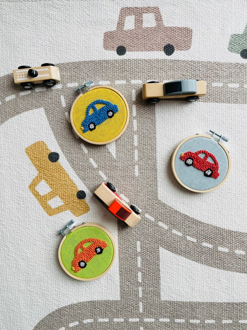 May include: Five wooden toy cars on a gray and white rug with a road design. Four of the cars are in embroidery hoops with colorful yarn. The cars are blue, red, orange, and red.