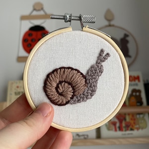 May include: A hand-stitched snail design in a wooden embroidery hoop. The snail has a brown shell and a grey body, set against white fabric. The hoop is secured with a metal screw and a wooden frame.