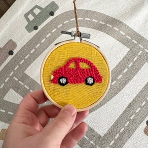 May include: A red car with black wheels and white accents is embroidered on a yellow fabric circle. The circle is framed with a wooden hoop.