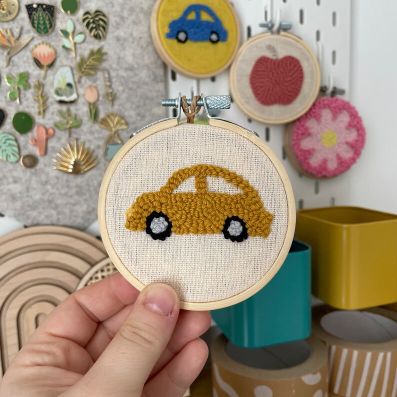 May include: A yellow car with black wheels and gray accents is embroidered on a white canvas in a wooden hoop. The hoop is being held by a hand.