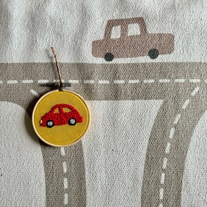 May include: A red car embroidered on a yellow felt circle, framed in wood. The circle is hanging from a string on a gray and white fabric background with a brown car design.
