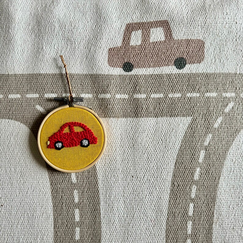 May include: A red car embroidered on a yellow felt circle, framed in wood. The circle is hanging from a string on a gray and white fabric background with a brown car design.