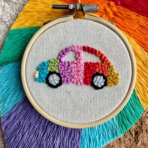 May include: A colorful embroidered car design on a white canvas in a wooden embroidery hoop. The car is made of rainbow colored yarn and has black wheels.