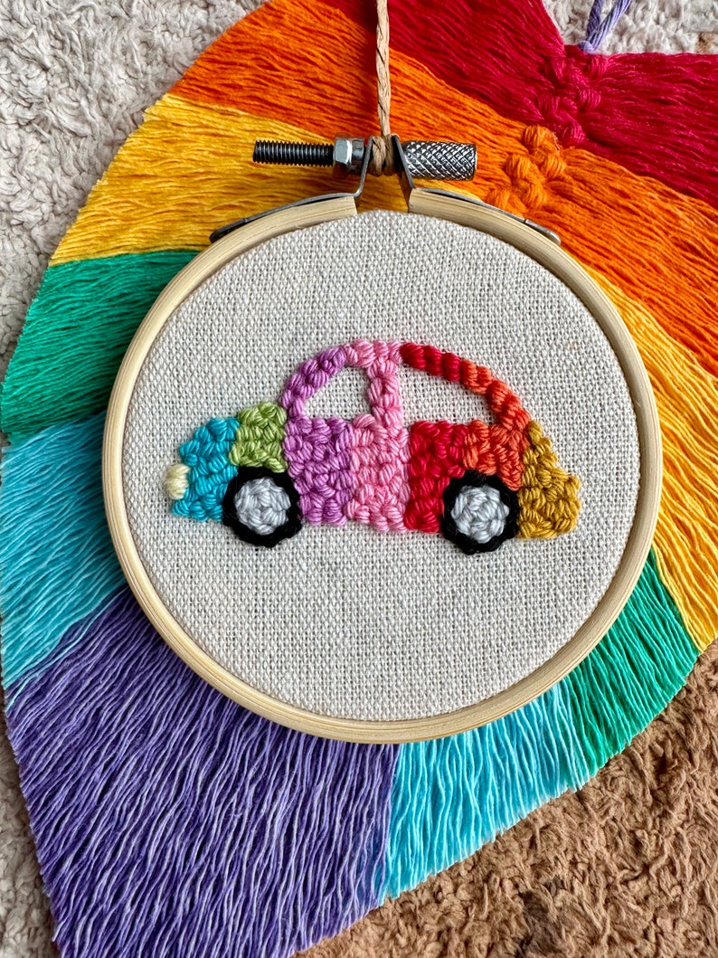 May include: A colorful embroidered car design on a white canvas in a wooden embroidery hoop. The car is made of rainbow colored yarn and has black wheels.