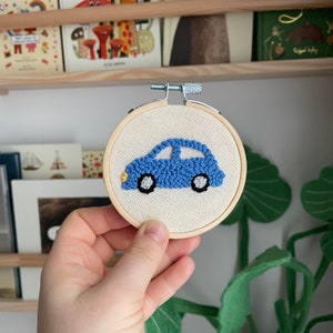 May include: A blue car embroidered on a white fabric stretched over a wooden embroidery hoop. The car has black wheels and yellow headlights.
