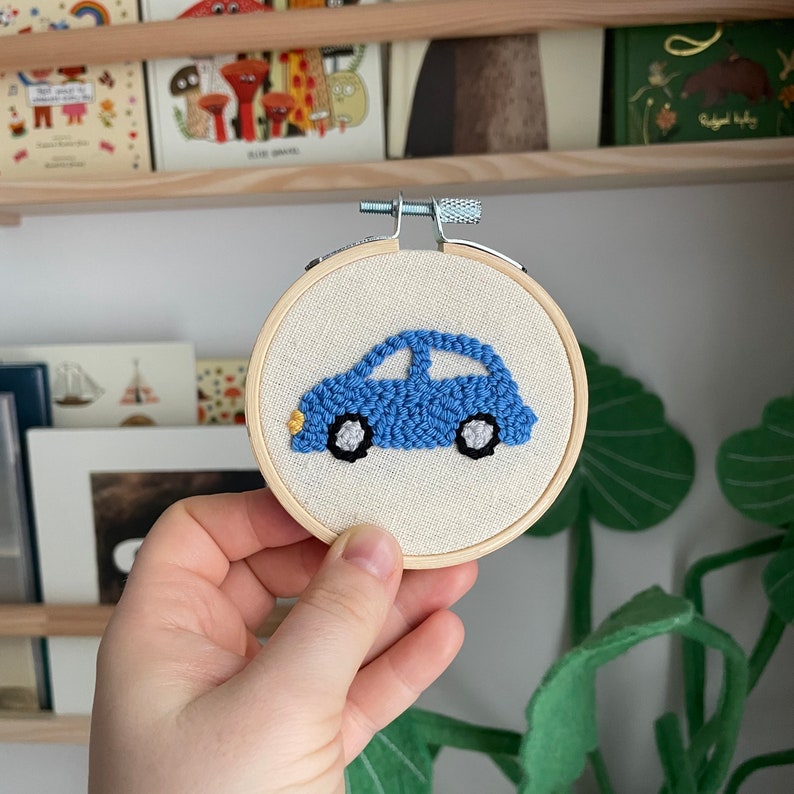May include: A blue car embroidered on a white fabric stretched over a wooden embroidery hoop. The car has black wheels and yellow headlights.