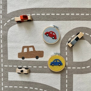 May include: A set of four wooden toy cars on a gray and white play mat. The cars are red, brown, blue, and gray. The cars are embroidered on felt circles.