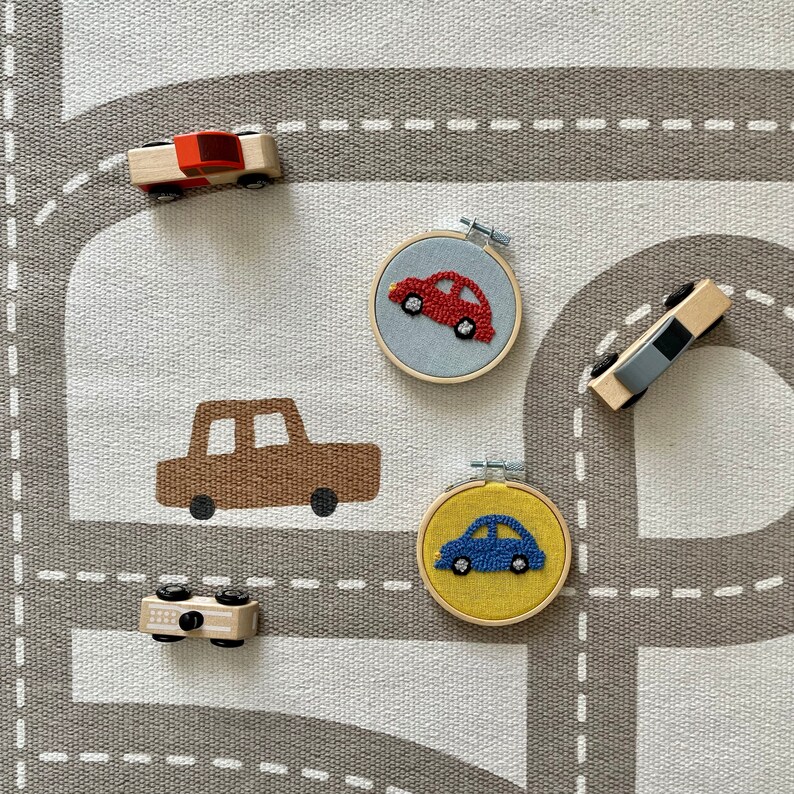 May include: A set of four wooden toy cars on a gray and white play mat. The cars are red, brown, blue, and gray. The cars are embroidered on felt circles.