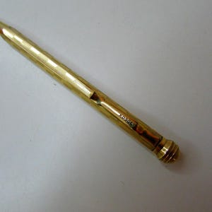 Könnte beinhalten: Ein goldfarbener mechanischer Bleistift im Vintage-Stil mit strukturierter Oberfläche und spitzer Spitze. Auf der Seite ist das Wort "KOSGA" eingraviert. Ein Schreibgerät.