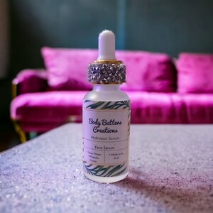 Może przedstawiać: Przezroczysta butelka szklana z złotą zakrętką i białą etykietą z napisem "Body Butters Creations Hydration Serum Face Serum Rose Water Manuka Honey Hyaluronic Acid".