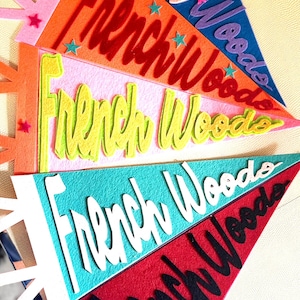 Könnte beinhalten: Eine Sammlung farbenfroher Filz-Wimpel mit dem Schriftzug "French Woods" in verschiedenen Schriftarten und Farben. Die Wimpel sind in Rosa, Orange, Blau und Rot gehalten und mit sternförmigen Verzierungen versehen.
