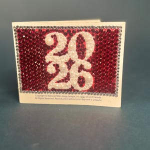 K&ouml;nnte beinhalten: Eine rechteckige Karte mit der Zahl 2026 aus wei&szlig;en Perlen auf einem Hintergrund aus roten Strasssteinen, umrandet von klaren Kristallen. Der untere Text lautet: Copyright National Mah Jongg League, Inc. US and Canada.