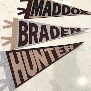 Könnte beinhalten: Drei Filz-Wimpel mit den Namen: Maddox, Braden und Hunter. Die Wimpel sind braun, grau und beige, mit den Namen in kontrastierenden Farben. Die Wimpel sind dreieckig, mit einem klassischen Schulbanner-Design.