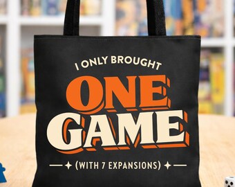 Op de afbeelding: Zwarte draagtas met de tekst "I ONLY BROUGHT ONE GAME (WITH 7 EXPANSIONS)" in oranje en wit. De tas heeft zwarte handvatten en staat op een houten oppervlak. Een blauw spelstuk en een witte dobbelsteen liggen ook op het oppervlak.