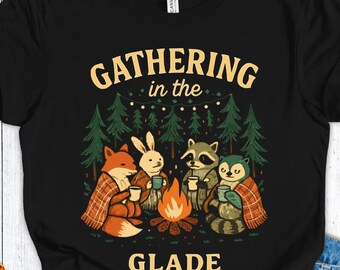 Op de afbeelding: Zwart t-shirt met een afbeelding van bosdieren rond een kampvuur. De vos, het konijn, de wasbeer en de uil zijn in dekens gewikkeld en houden bekers vast. De tekst "GATHERING in the GLADE" staat boven en onder de illustratie.