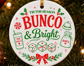Op de afbeelding: Een witte keramische kerstversiering met de woorden "Bunco & Bright" in rood en groen. De versiering heeft dobbelstenen, een bel, een drankje en een cadeauzakje, allemaal in een feestelijk ontwerp. De versiering hangt aan een kerstboom.
