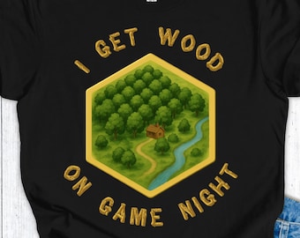 Op de afbeelding: Zwart t-shirt met een goudkleurige zeshoekige afbeelding. De afbeelding toont een boslandschap met een rivier en een klein huis. De tekst "I GET WOOD ON GAME NIGHT" staat in een boog rond de afbeelding.