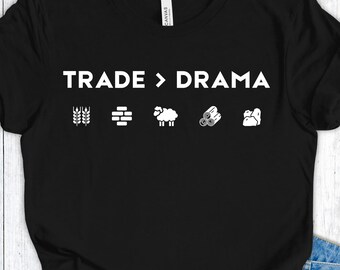 Op de afbeelding: Zwart t-shirt met de witte tekst "TRADE > DRAMA" en iconen van tarwe, een gebouw, een schaap, boomstammen en een berg. Het shirt is gemaakt van een zacht materiaal.