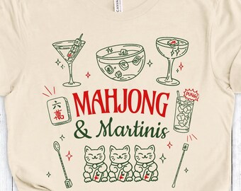 Op de afbeelding: Crèmekleurig T-shirt met een groen en rood grafisch ontwerp. Het ontwerp bevat martiniglazen, een kom en de tekst "MAHJONG & Martinis". Daaronder staan drie illustraties van gelukskatten.