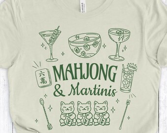 Op de afbeelding: Lichtbeige t-shirt met een groen grafisch ontwerp. Het ontwerp bevat illustraties van martini glazen, mahjong tegels en drie katten. De tekst "MAHJONG & Martinis" is ook aanwezig.