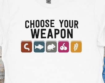 Op de afbeelding: Wit t-shirt met de zwarte tekst "CHOOSE YOUR WEAPON". Daaronder vijf gekleurde vierkanten met afbeeldingen: een worm, een vis, een muis, kersen en tarwe.