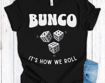 Op de afbeelding: Zwart T-shirt met het woord "BUNCO" in witte letters boven drie dobbelstenen. De zin "IT'S HOW WE ROLL" staat eronder. Het shirt is gemaakt van een zacht materiaal, geschikt voor casual wear.