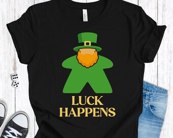 Op de afbeelding: Zwart T-shirt met een groene kaboutervormige figuur, een oranje baard en een groene hoed. De gouden tekst eronder luidt "LUCK HAPPENS". Het T-shirt wordt gedragen met een blauwe spijkerbroek en zwarte sneakers.