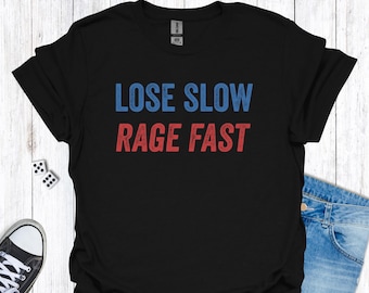 Op de afbeelding: Zwart t-shirt met de tekst "LOSE SLOW RAGE FAST" in blauwe en rode letters. Het shirt wordt gecombineerd met een spijkerbroek en dobbelstenen. Een casual t-shirt voor dagelijks gebruik.