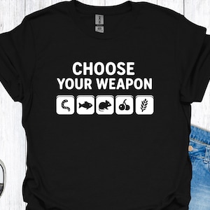 Peut inclure: T-shirt noir avec l'inscription blanche "CHOOSE YOUR WEAPON". En dessous, quatre icônes blanches : un ver, un poisson, une souris, des cerises et un épi de blé. Le t-shirt est porté avec un jean bleu et des baskets.
